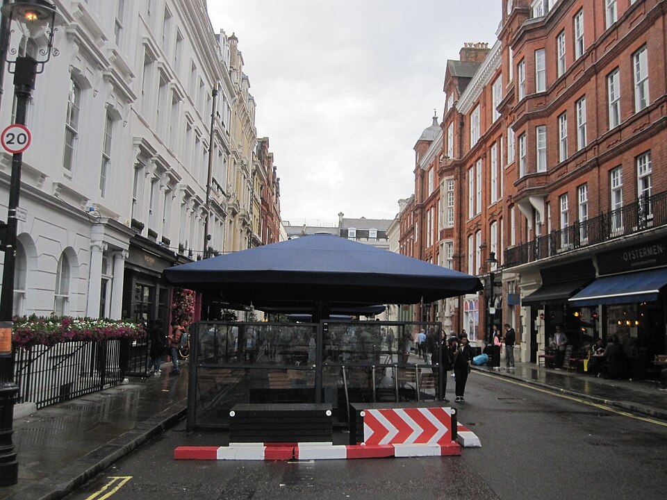 Henrietta Street on a wet day, white stucco along the left and red brick mansion flats along the right, with an awning set out in the middle of the road.