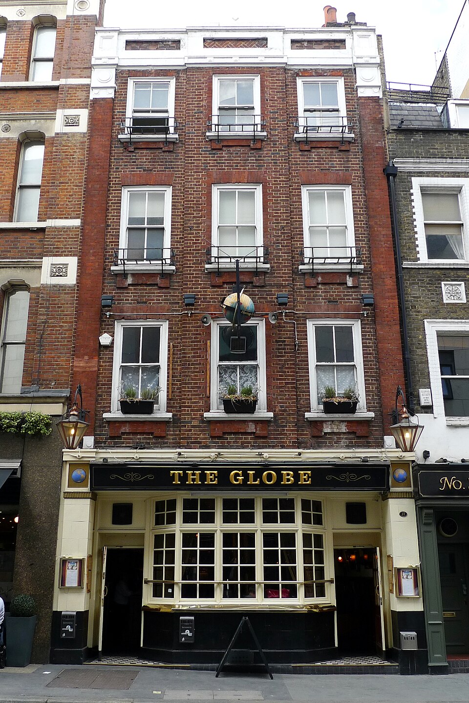 The brick front of 37 Bow Street with a black and gold pub fascia across the ground floor, three floors of sash windows with window boxes above it and a lamp on either side of the door.