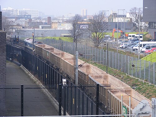 A stone train of empty wagons on the line at Hackney Wick, seen from above, with HACKNEY WICK painted across a warehouse wall beyond the track.