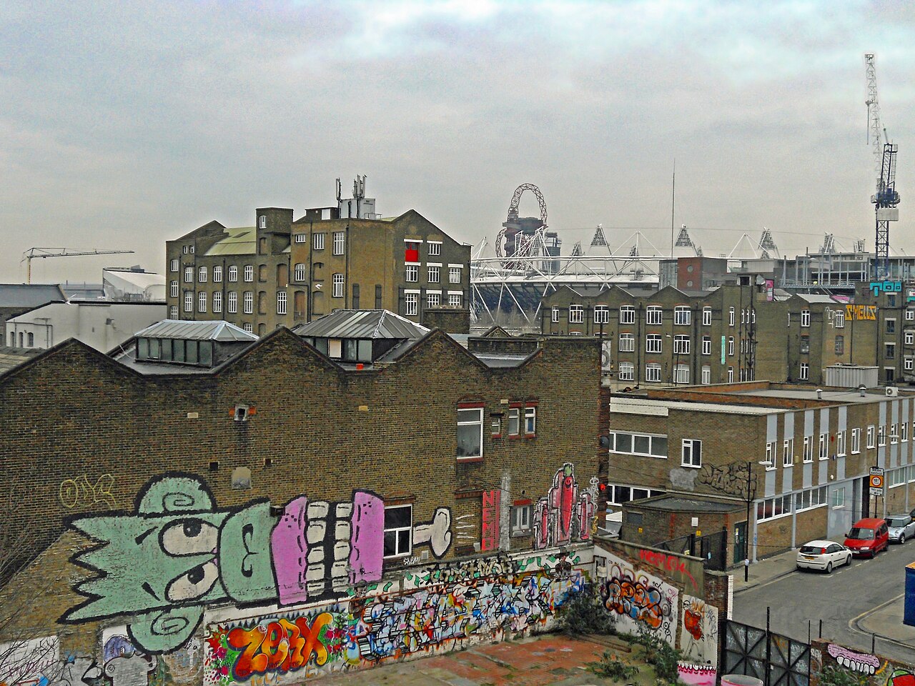 The rooftops of Hackney Wick from an upper window, brick works with skylights along the ridge, a side street with parked cars, and the Olympic Stadium and the Orbit behind them.