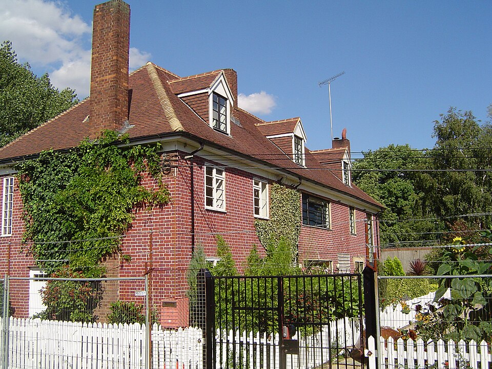 The lock keepers' cottages at Old Ford Lock in 2005, a red brick house with three dormers, a tall chimney and ivy up the gable, behind a white picket fence.