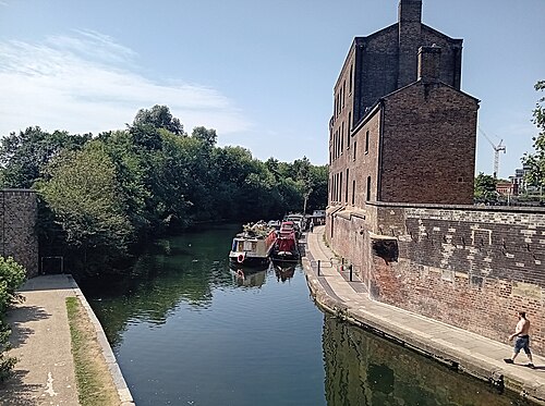 The Regent's Canal at King's Cross, a brick warehouse rising straight out of the water on the right and narrowboats moored along the far bank.