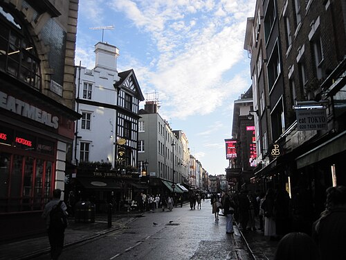 Old Compton Street on a wet afternoon, a mock Tudor pub corner on the left and lit signs down the right, people out on both pavements.