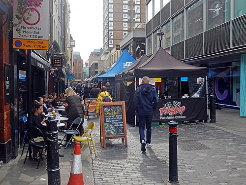 Berwick Street market, stalls under square gazebos down the middle of the road, cafe tables on the left and a no vehicles sign at the corner.