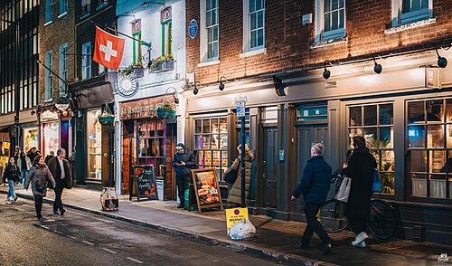 Wardour Street at night, a lit brick terrace of restaurants with a Swiss flag hung over one of them and people along the pavement in front.