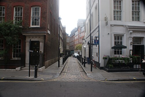 Meard Street seen from Dean Street, a single lane of granite setts with bollards along both kerbs, red brick and iron railings on the corner to the left and white stucco to the right.