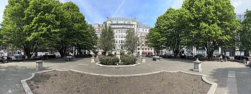 Golden Square from inside the garden, trees down both sides and a stone statue on a plinth in the middle of a round flower bed, with the buildings of the square behind.