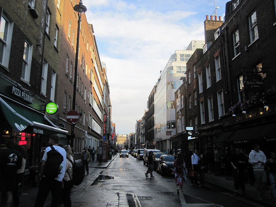 Dean Street in the evening, brick fronts four and five storeys with restaurants at street level, a pub with a hanging sign on the right and a Soho Theatre sign further down the block.