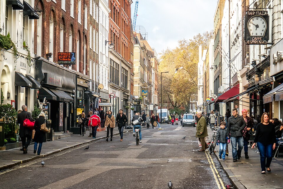 Frith Street looking north toward the trees of Soho Square, the Ronnie Scott's sign on the left and the Bar Italia clock on the right, four and five storey fronts along both sides.