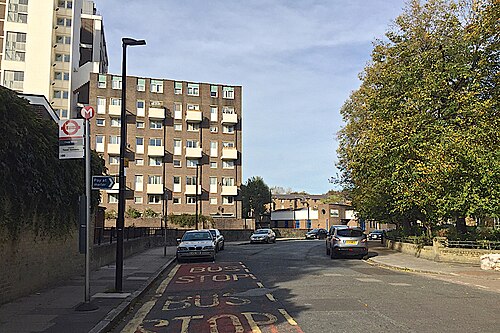 Prusom Street with a brick and concrete block of flats on the left, a bus stop flag on the pavement and the bus stop markings painted twice over on the road.