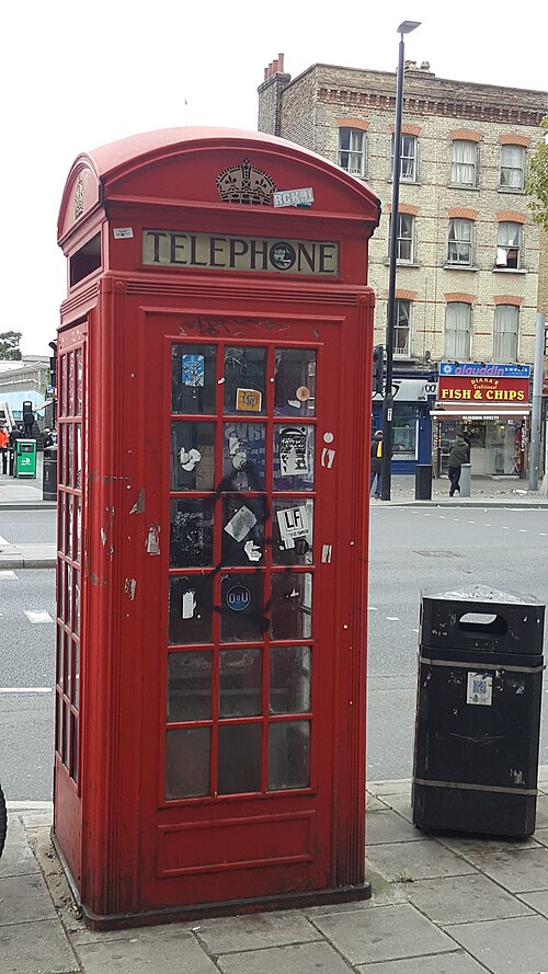 A K2 telephone kiosk on the pavement outside the Royal London Hospital, with a sweet shop and a fish and chip shop in the terrace across Whitechapel Road.