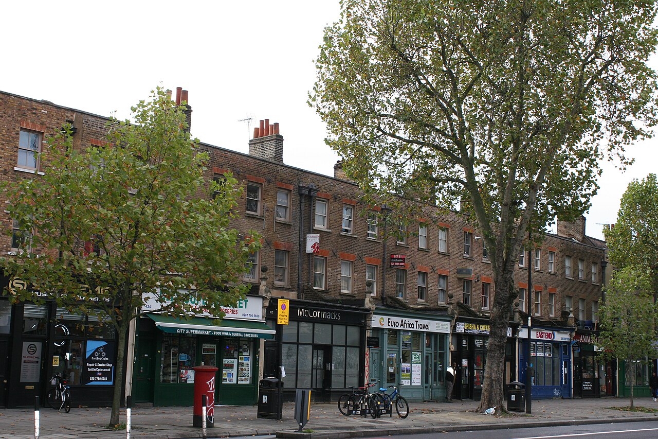 The terrace at 90 to 126 Mile End Road, stock brick houses over shops with plane trees along the pavement and a pillar box at the kerb.