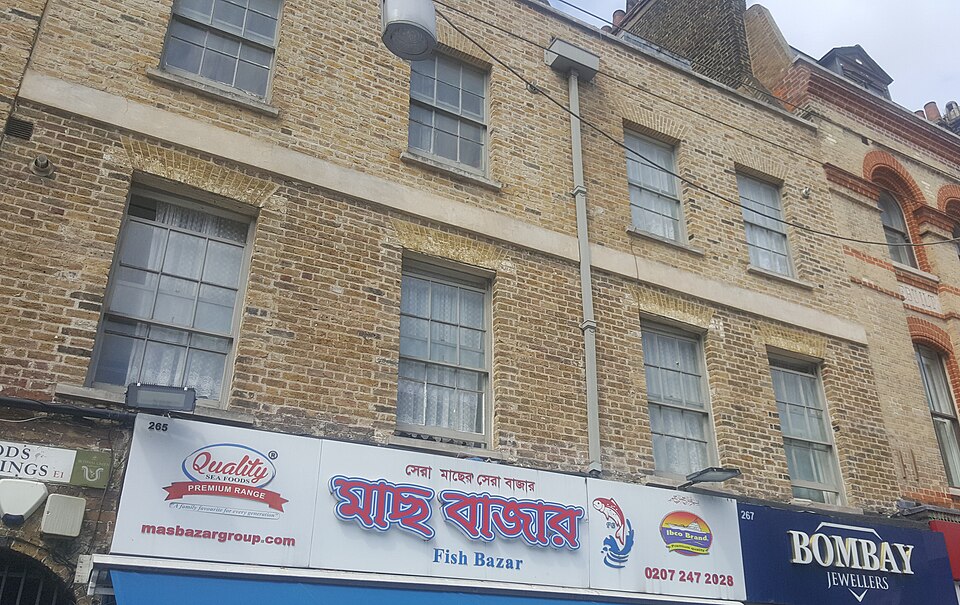 The upper floors of 265 and 267 Whitechapel Road in stock brick, with the fish shop sign in Bengali below and the Wood's Buildings sign at the left edge.
