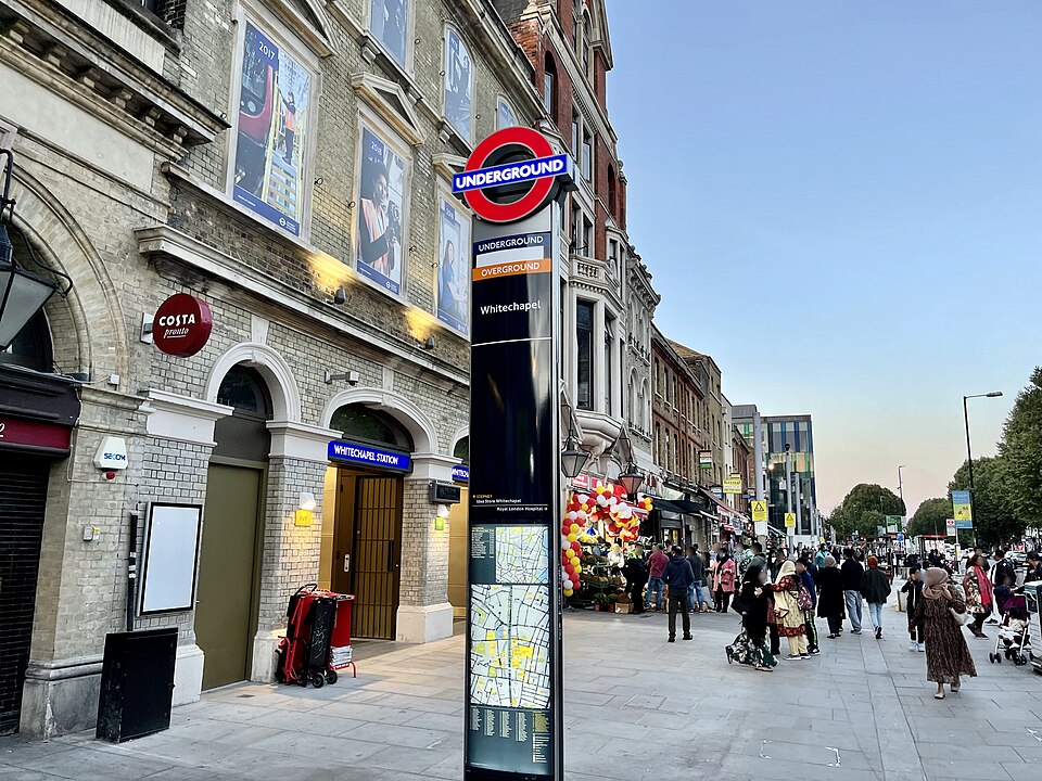 The Whitechapel Road entrance to Whitechapel station in the evening, with the Underground and Overground totem on the pavement and the market stalls beyond it.