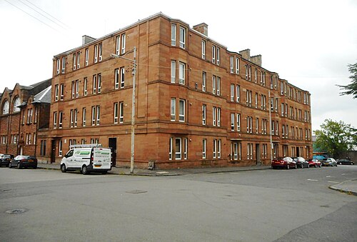 A red sandstone tenement on the corner of Blackie Street, with a blank rendered gable on the near side and cars parked along both sides.