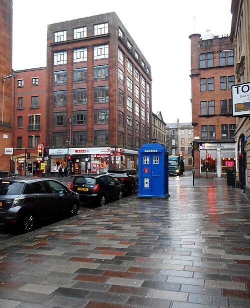 A blue police box on the paved part of Wilson Street after rain, with a red sandstone block behind it and a Spar and a post office at street level.