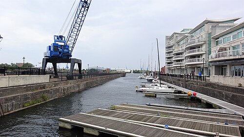 A preserved blue portal crane beside the stone walled Mount Stuart Graving Dock, modern apartment blocks behind.