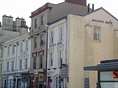 A row of painted stucco three and four storey buildings on Bute Crescent, hotel and pub signage across their ground floors.