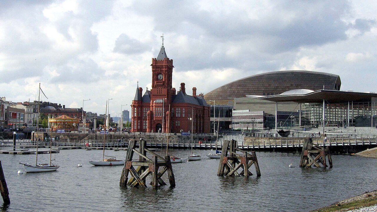 A wide view across the water showing the red brick Pierhead Building, the Senedd's canopy roof and the bronze shell of the Wales Millennium Centre together, with sailing boats moored at wooden pontoons in front.