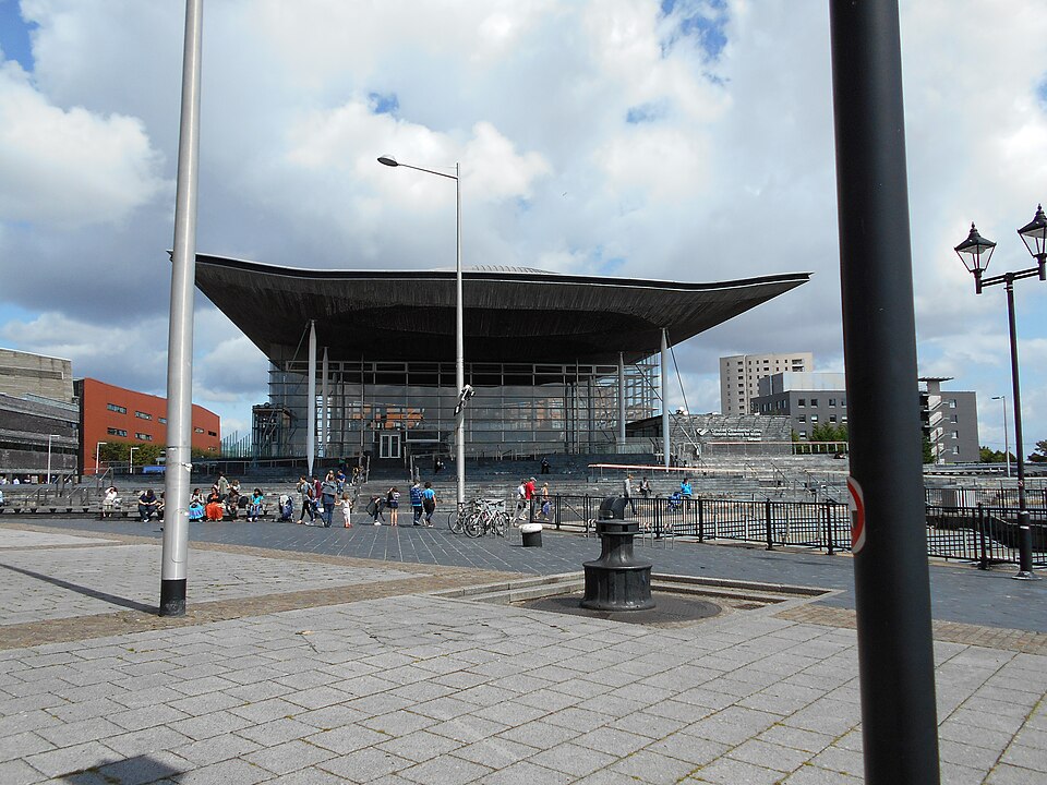 The Senedd's swooping roof canopy on slender white columns, glass walls beneath, with the National Assembly for Wales sign at the entrance.