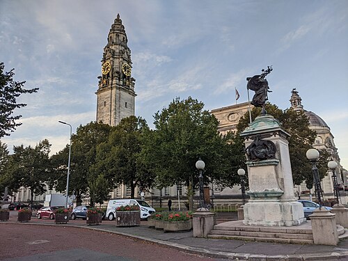 Cardiff City Hall's clock tower and domed corner rising behind trees and lamp posts, the bronze winged figure of the South African War Memorial on its stone plinth in the foreground.