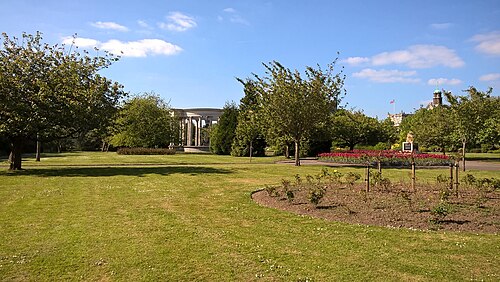 A lawn in Alexandra Gardens with flowerbeds and trees, the Welsh National War Memorial's circular stone colonnade in the middle distance and a dome visible beyond it.