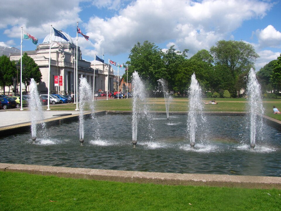 The National Museum Cardiff's colonnaded portico and dome behind a row of flagpoles and fountains, cars parked along the forecourt.