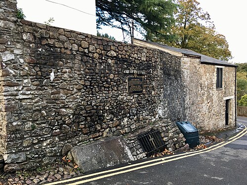 A rubble stone wall with a plaque marking St Teilo's Well runs beside a bending lane with double yellow lines, a slate roofed outbuilding beyond it.
