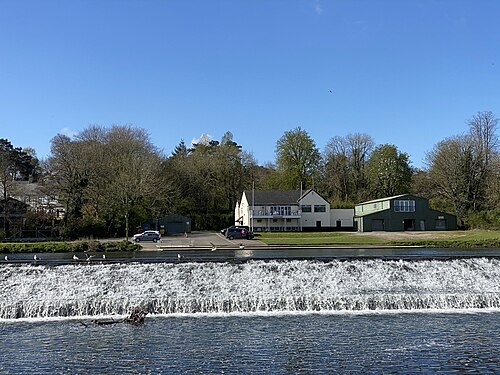 Llandaff Rowing Club's white clubhouse and a green boat shed stand behind a weir on the River Taff, white water pouring over the sill.