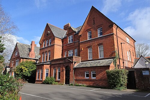 Howells School's Victorian sixth form building in red brick with gabled dormers and bay windows, set back from the road behind a car park.