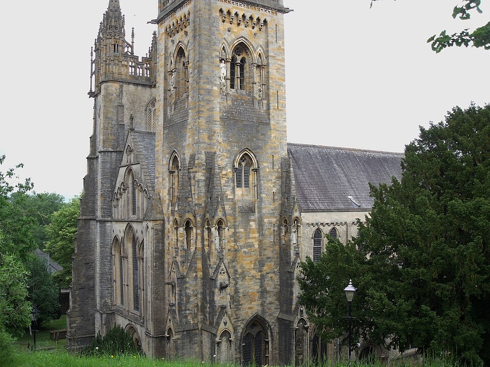 The cathedral's west towers in buff and grey stone, arcaded and pinnacled, with tall lancet windows above the churchyard grass and trees.