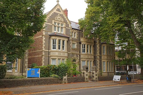 A pair of Cathedral Road villas in rock faced stone, a central gable and arched doorway, a mature tree between them and a charity sign at the railings.