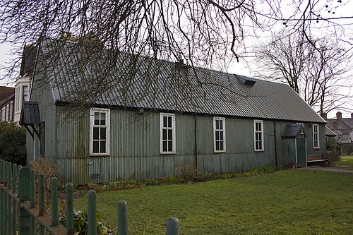 A plain corrugated iron church hall painted green, arched sash windows and a small door, seen from behind iron railings.