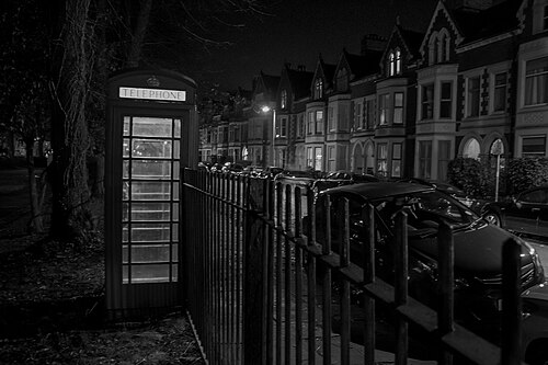 A red telephone box beside iron railings at night, a crescent of gabled bay fronted terraces lit up along the street behind it.