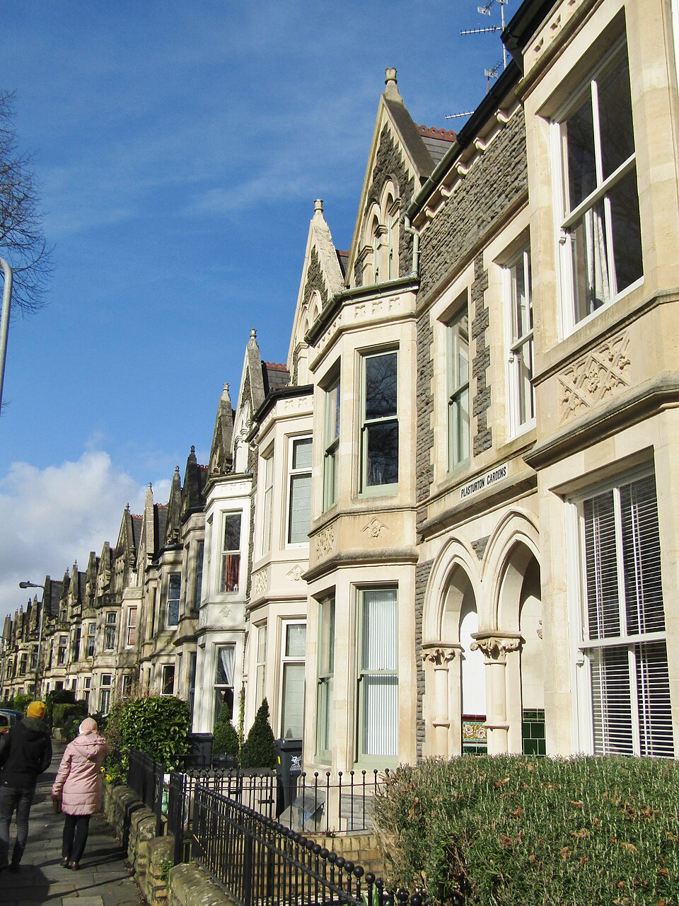 A gothic revival stone terrace on Plasturton Gardens, the name cut into the corner stone above an arched doorway, two people walking away down the pavement.