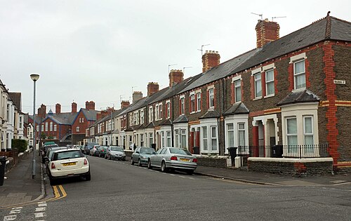 Another straight terraced street, dark slate roofs and a run of brick chimney stacks over bay fronted houses, a pale blue school building visible at the far end.