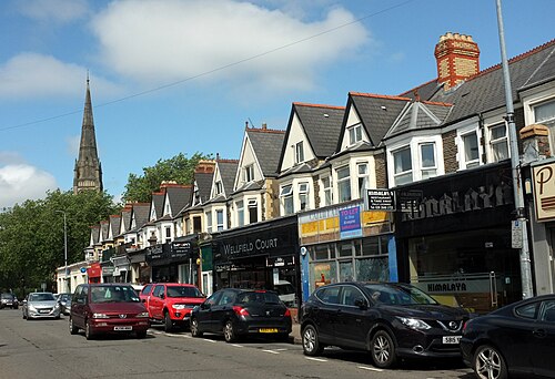 Wellfield Road's shopfronts under gabled bay windowed flats, a church spire rising behind the roofline, cars parked nose to tail along the kerb.