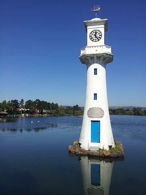 The white lighthouse memorial standing on its own rock base in the lake, a ship weathervane and a working clock built into the tower, a blue door at its foot.