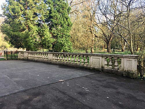 A stone balustrade bridge with urn shaped balusters between the lake and the conservatory, evergreen and bare trees behind it.
