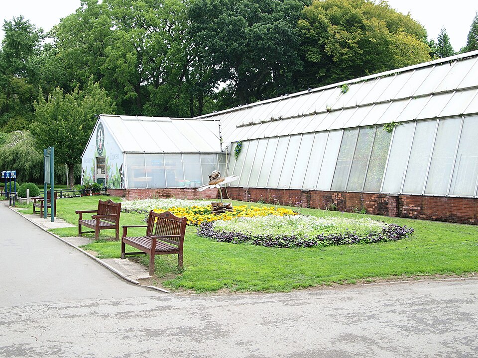 The long glass and brick conservatory in Roath Park's gardens, two benches and a circular flower bed with a bird sculpture fountain on the lawn in front of it.