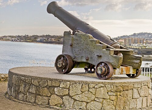 A historic iron cannon on a wooden carriage, mounted on a circular granite plinth near the sea.
