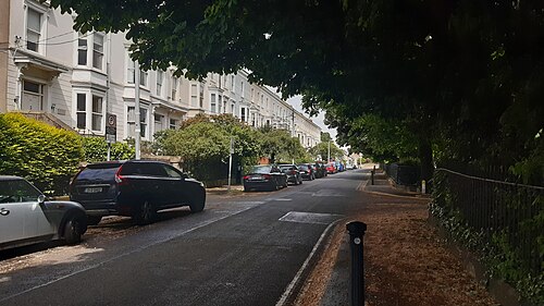 A tree lined residential street bordered by a continuous white stucco terrace, with bay windows and parked cars.