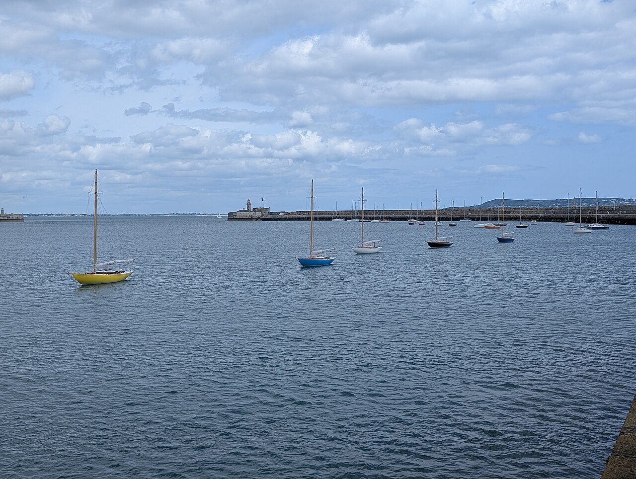 Sailing yachts moored in harbour waters enclosed by the two long stone pier breakwaters, under a cloudy sky.