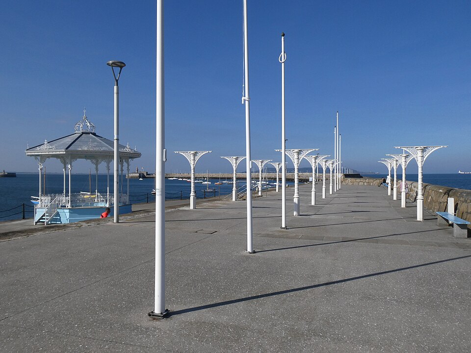 A wide concrete pier with the light blue Victorian bandstand, white cast iron shelter supports and white flagpoles beside open sea.