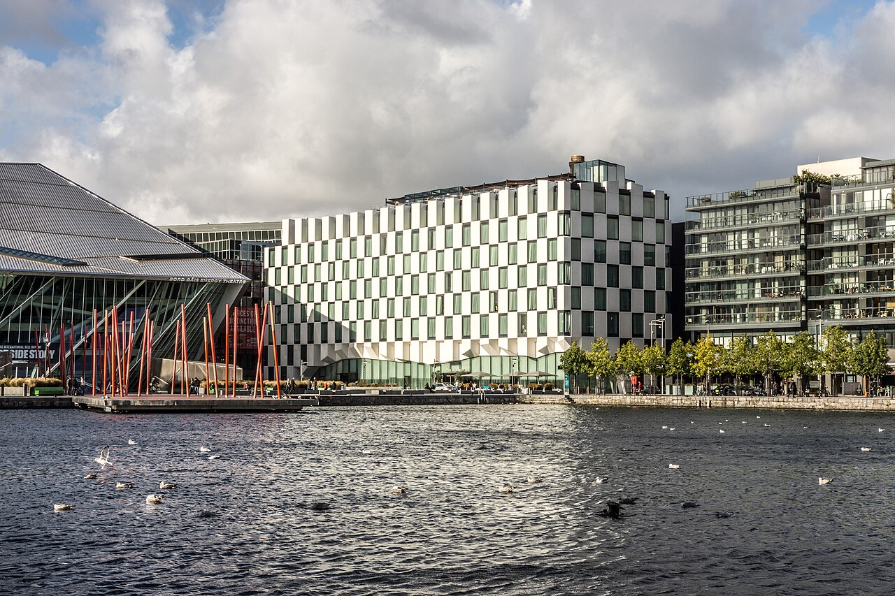 The Marker Hotel's black and white checkerboard facade beside the Bord Gais Energy Theatre's folded roofline, seen across the dock basin with gulls on the water.