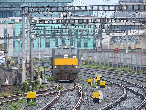 A train arriving on the tracks below Grand Canal Dock, glass office towers and the station's arched roof behind it.