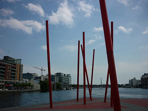 The red poles at Grand Canal Square close up, with apartment blocks and a construction crane on the far side of the water.