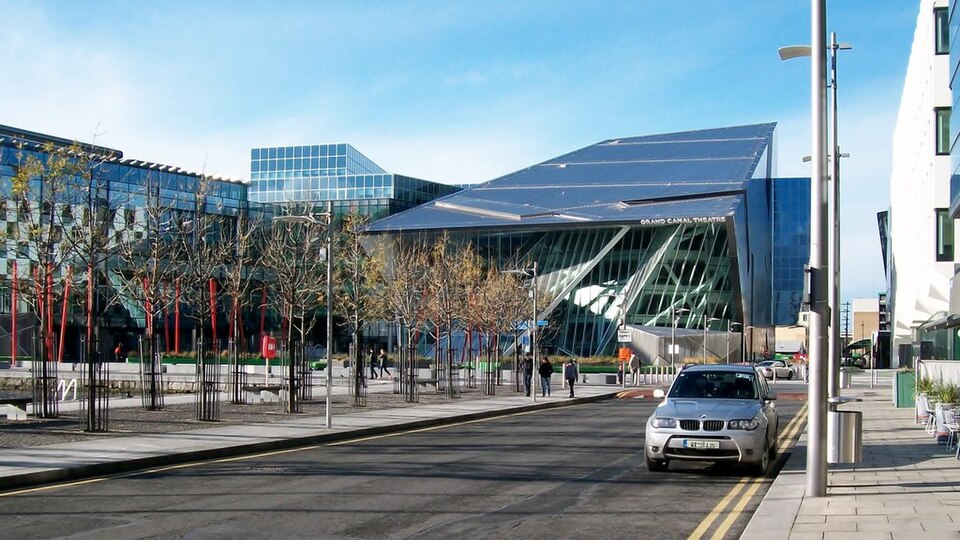 The Grand Canal Theatre entrance from the square, a row of red poles in front, trees along the paving and the theatre's angled glass front behind.