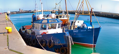 Blue fishing trawlers moored along the harbour pier, yellow bollards on the quay and a stone lighthouse in the background.