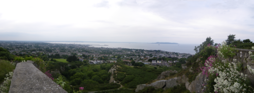 The headland seen from an elevated stone wall on the cliff path, green paths and houses above the coastline and Dublin Bay beyond.